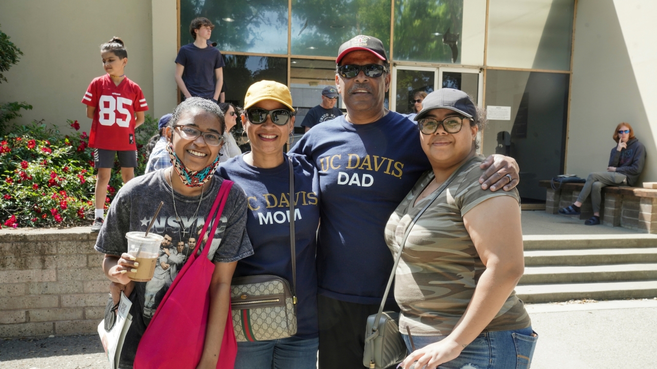Four people stood in front of a building. The woman second from the left is wearing a UC Davis Mom T-Shirt, and the man to the right of her is wearing a UC Davis Dad T-Shirt.