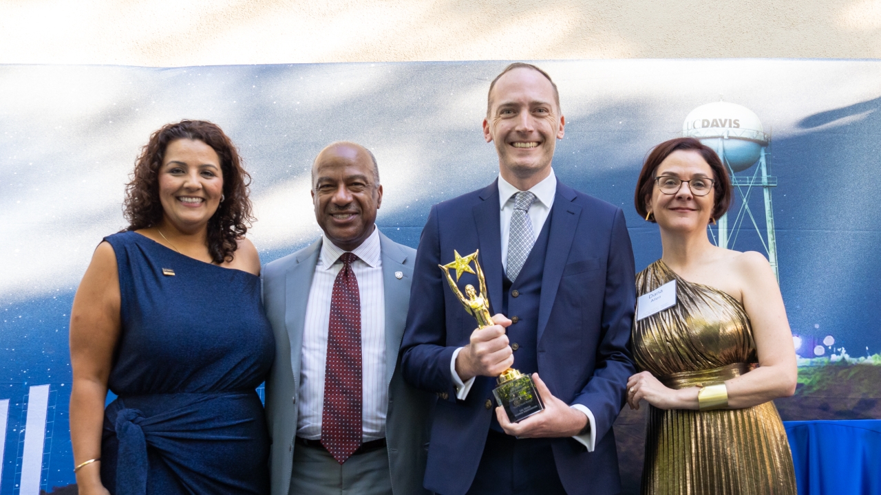 Four people on stage at the awards ceremony. Charles Melton in the middle holds the gold statue.