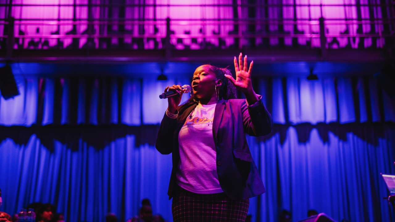 Bland holding a microphone and animatedly reciting poetry, holding one hand up, in a dark theater with blue and purple lights in the background