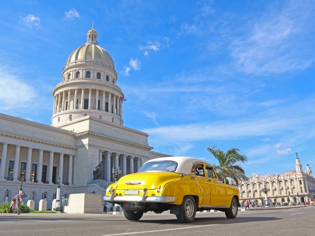 A classic yellow car drives past a grand building under a bright blue sky.