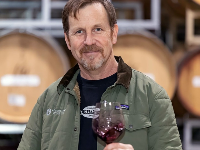 Smiling man in green jacket holding red wine glass in cellar with oak barrels