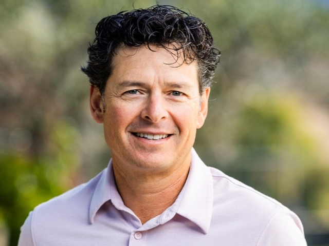 Smiling middle-aged man with dark curly hair in a light shirt, blurred outdoor background