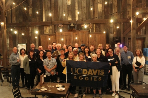 A group of alumni and UC Davis staff posing at a wine club release party at a rustic winery in the evening with string lights.