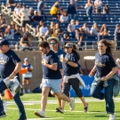 In the forefront, 8 people are in the motion of running on the football field in Aggie Pride blue shirts. In the background are people standing on the sidelines of the field and you can also see fans sitting the stands.