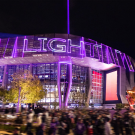 Modern arena facade lit purple at night, blurred crowd and city skyline behind