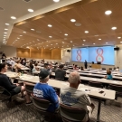 Conference lecture hall with attendees facing large screen displaying two red 8s