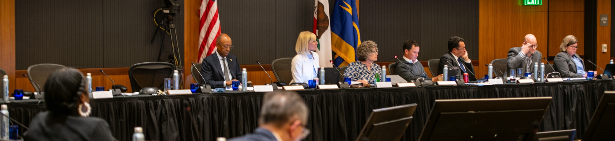A group of people seated at a long table in a lecture hall attending a UC Regents meeting. There are flags rolled up between meeting participants and meeting participants are seated behind microphones.